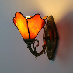 An orange Tiffany-style wall sconce with a flower-shaped stained glass shade, illuminated and mounted on a distressed gold metal arm against a textured wall.