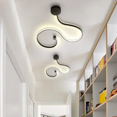 Two black wave-shaped LED ceiling lights with warm light mounted on a white ceiling in a hallway with bookshelves.