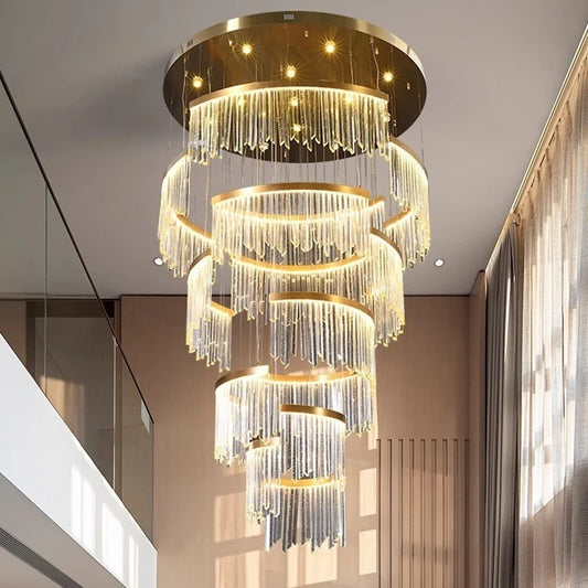A grand, multi-tiered gold chandelier with cascading clear crystal rods, illuminated with warm light, installed in a modern, high-ceiling foyer with marble accents and dual staircases.