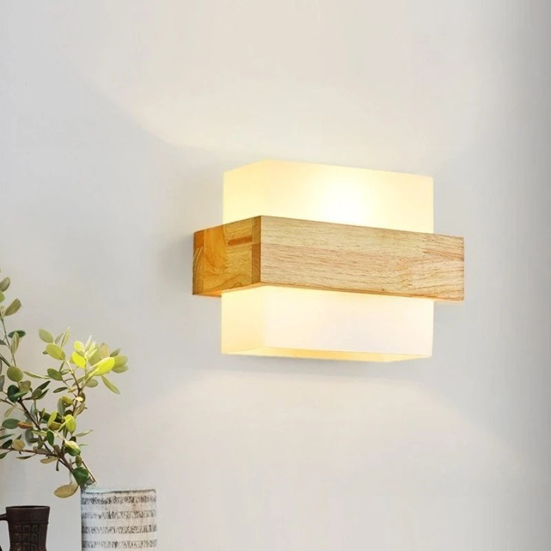 A wood and glass LED wall sconce mounted on a white wall, positioned to the right of a vase with green foliage.