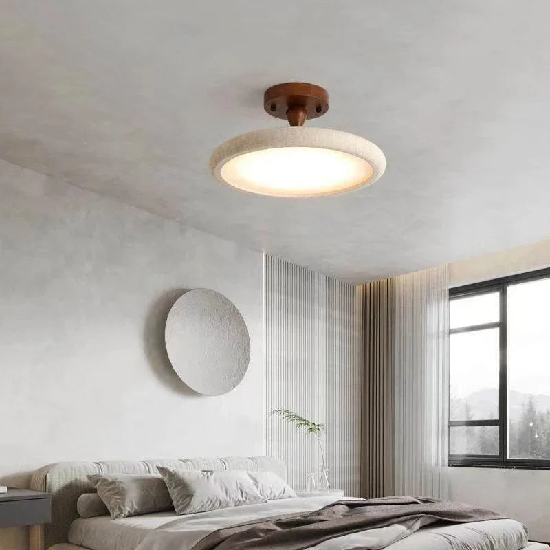 A round ceiling light fixture with a stone-like textured rim and a dark wood base, illuminated and mounted on a white textured ceiling above a bed.