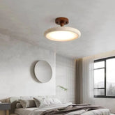 A round ceiling light fixture with a stone-like textured rim and a dark wood base, illuminated and mounted on a white textured ceiling above a bed.