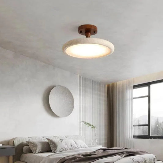 A round ceiling light fixture with a stone-like textured rim and a dark wood base, illuminated and mounted on a white textured ceiling above a bed.