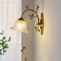 Side profile of a gold floral wall sconce with a glass shade, showcasing its curved arm and leaf and rose embellishments against a white wall.