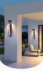 A modern black wall sconce with a natural stone insert mounted on a white outdoor wall, illuminated at dusk.