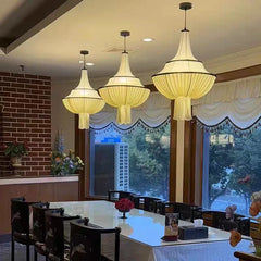 Three white fabric pleated pendant lights with black trim hanging over a dining table with chairs, near a window with curtains.