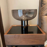 A table lamp with a smoked gray glass bowl shade and a dark metal base, placed on a wooden nightstand.