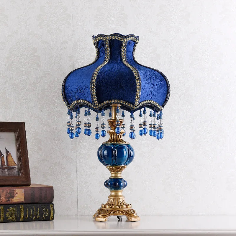 Front view of a table lamp with a blue fabric shade, gold trim, and dangling beads, set on a golden base with blue accents.