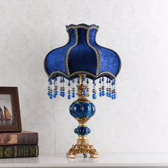 Front view of a table lamp with a blue fabric shade, gold trim, and dangling beads, set on a golden base with blue accents.