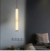 A long, slender marble cylinder pendant light with natural stone inclusions hanging in a dimly lit room next to a bed.