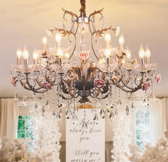 Detail shot of the chandelier's arm, highlighting the texture of the gold finish, the delicate pink ceramic rose, and a sparkling teardrop crystal.