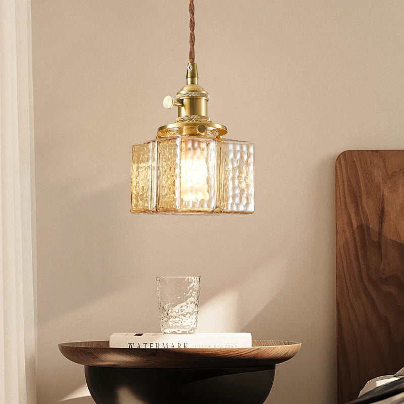A single glass square pendant light with a brass fixture hanging above a small wooden table with a glass.
