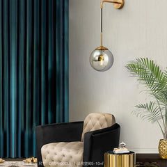A gold wall sconce with a clear glass globe mounted on a textured wall next to a dark armchair and teal curtains.