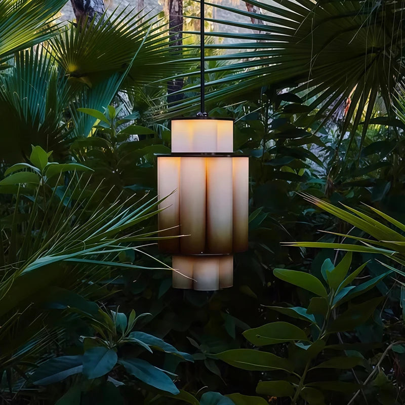A gold and white acrylic tiered pendant light hanging outdoors surrounded by lush green foliage.