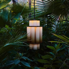 A gold and white acrylic tiered pendant light hanging outdoors surrounded by lush green foliage.