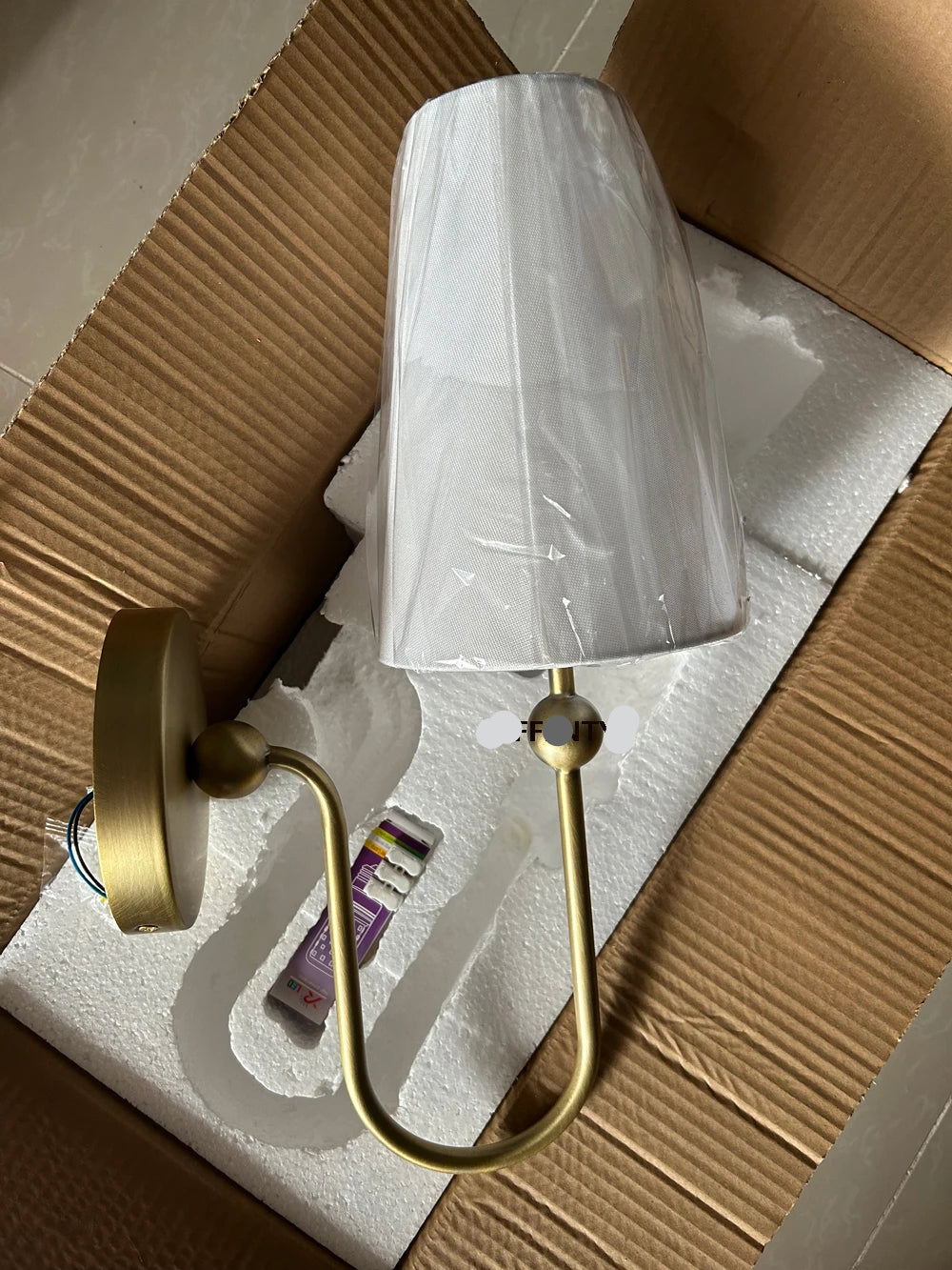 Wall-mounted light fixture with gold finish and white shade on a cardboard box.