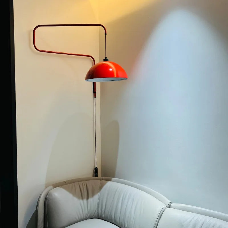 Red folding retractable wall lamp with a dome shade mounted on a light-colored wall above a white sofa, casting a warm light.