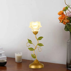 A gold table lamp with a flower-shaped glass shade and green leaves on a stem, placed on a wooden surface next to a candle and a vase of flowers.
