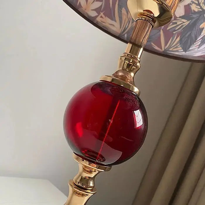 Close-up view of the round red glass orb accent on the polished brass stem of a table lamp, with a floral fabric shade visible above.