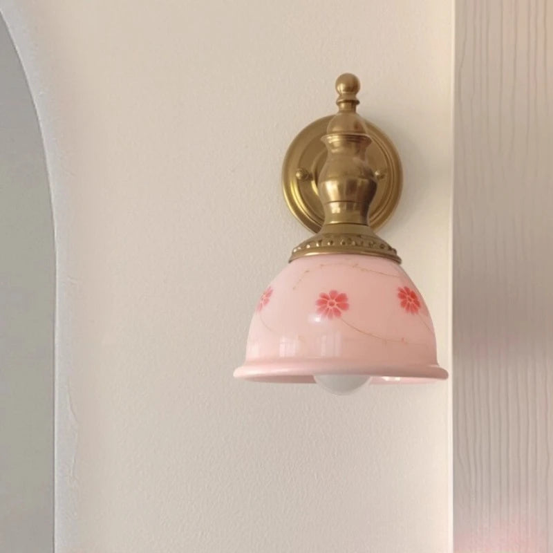 Wall-mounted light fixture with pink glass shade and gold metal fixture on a neutral wall.