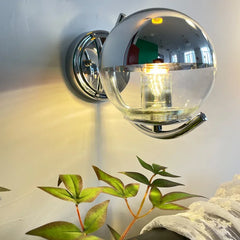 Wall-mounted light fixture with clear glass shade and metallic finish, illuminated against a neutral background.