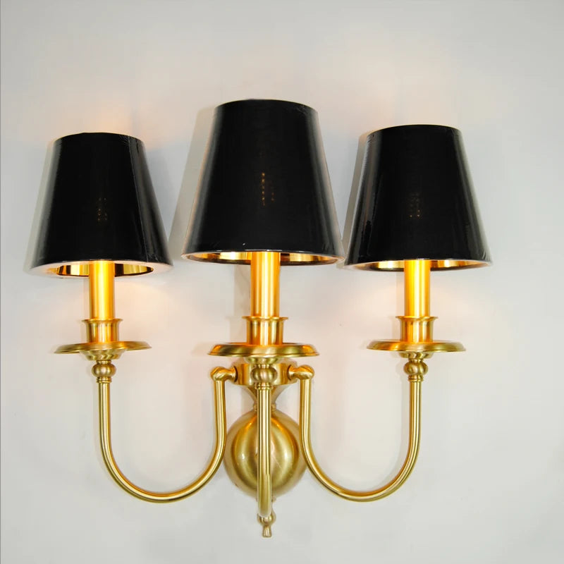 Three-arm wall sconce with black lampshades and gold metal finish on a white background