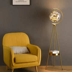 Yellow armchair with a cushion next to a decorative floor lamp against a grey wall. Modern glass ball LED tripod lamp for bedroom or study.