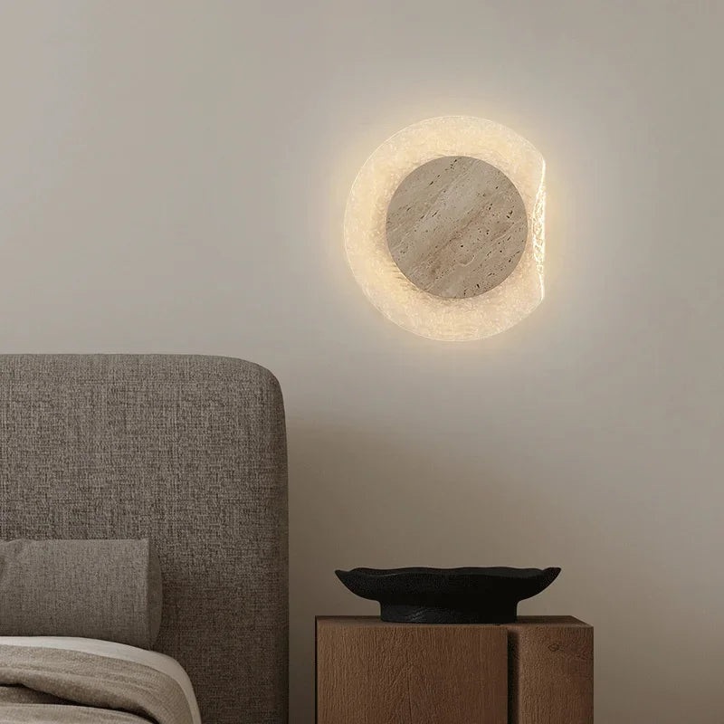 Round wall light fixture on a neutral wall above a bed with a wooden nightstand.
