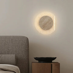 Round wall light fixture on a neutral wall above a bed with a wooden nightstand.