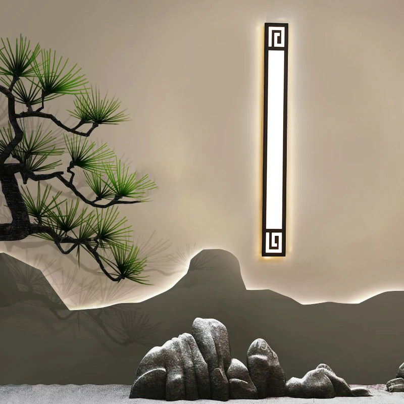 A single black geometric LED wall sconce mounted on a neutral wall, set against a backdrop of stylized rocks and a pine tree.