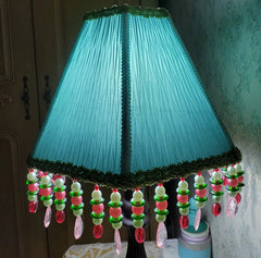 A light green pleated fabric empire lampshade with beaded tassels, illuminated from within, casting a soft glow.