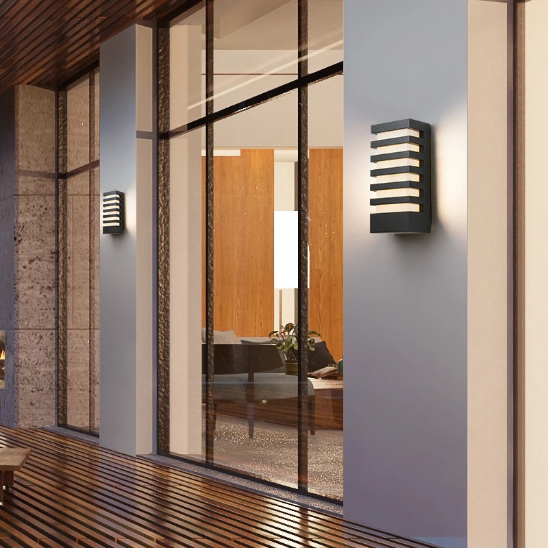 Two black modern outdoor wall sconces with horizontal light slats, mounted on a light-colored wall next to a large glass window showing an interior.