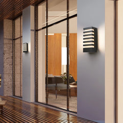 Two black modern outdoor wall sconces with horizontal light slats, mounted on a light-colored wall next to a large glass window showing an interior.
