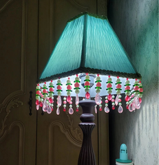 A teal blue pleated fabric empire lampshade with beaded tassels, fitted onto a lamp base in a dimly lit room.