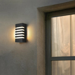 A close-up of a black modern outdoor wall sconce with horizontal light slats, mounted on a textured wall next to a window with sheer curtains.