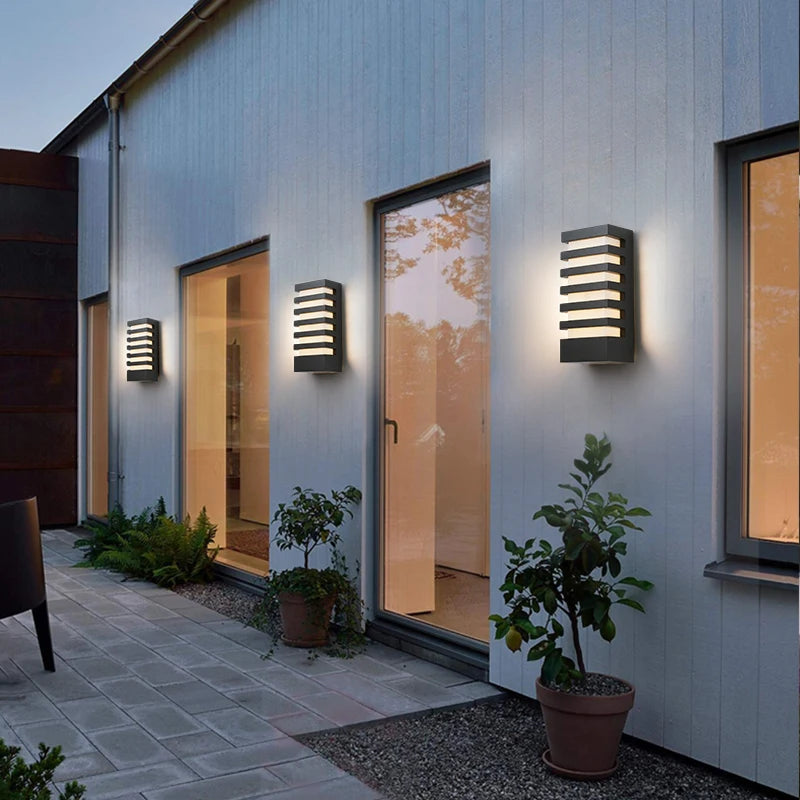 Three black modern outdoor wall sconces with horizontal light slats, mounted on a light-colored wall along a building facade with windows and plants.