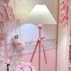 Side view of a pink floor lamp with a fabric shade and bow, next to a white wooden chair with a decorative pillow.