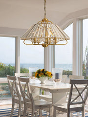 A gold crystal faceted pendant light suspended above a white dining table by a large window overlooking the ocean.