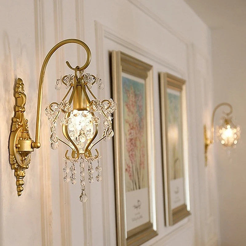 Two gold crystal wall sconces flank framed artwork on a white wall, demonstrating their decorative potential in a hallway or living space.
