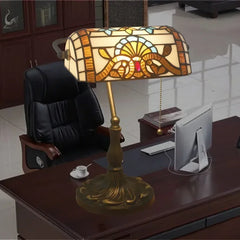 A Tiffany-style stained glass table lamp with an amber and blue patterned shade, set on a wooden desk in an office environment.