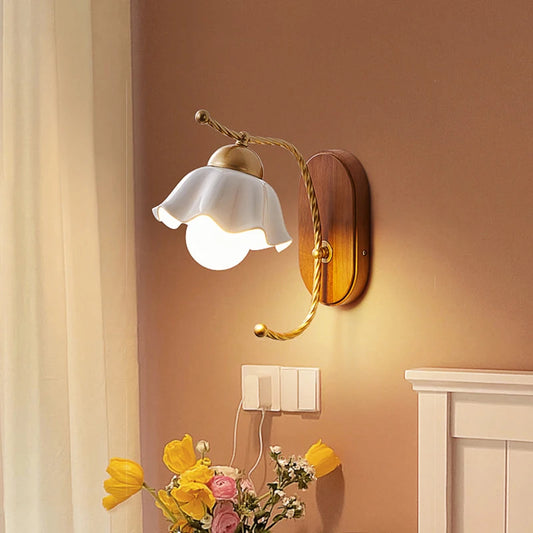 A wall sconce with a white ceramic flower shade and gold arm mounted on a wooden base, illuminated in a room with a brown wall and yellow flowers in the foreground.