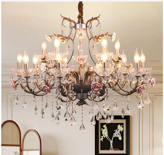 Front view of a decorative gold chandelier featuring intricate metalwork, pink ceramic roses, and clear crystal droplets, illuminating a room.