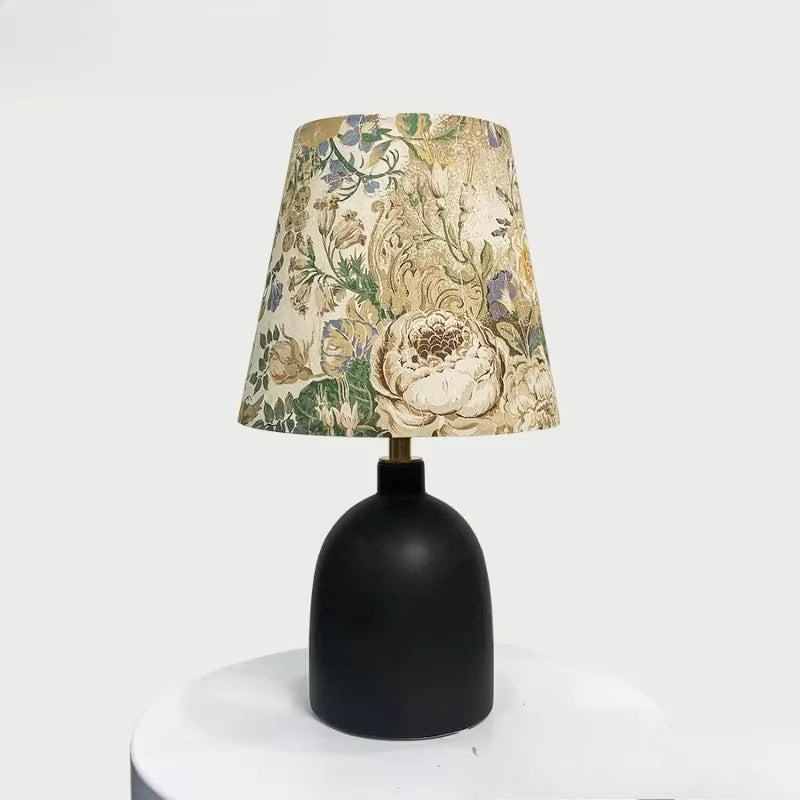 Black ceramic table lamp with a conical fabric shade featuring a vintage floral pattern, set on a white surface.
