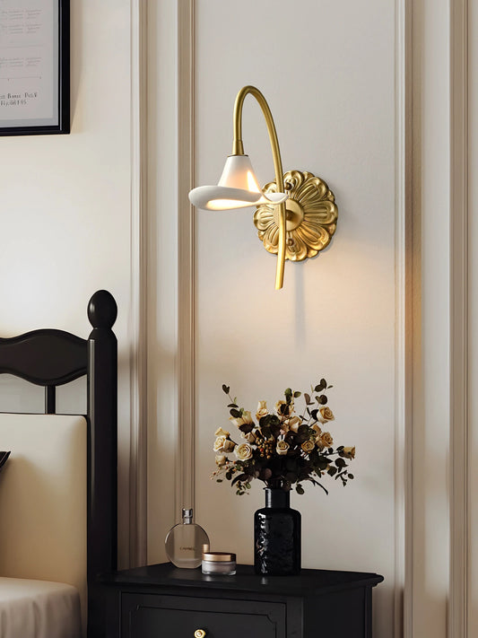 A gold floral ceramic wall sconce illuminated and mounted on a wall above a bedside table with a vase of flowers and a perfume bottle.