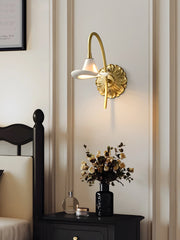 A gold floral ceramic wall sconce illuminated and mounted on a wall above a bedside table with a vase of flowers and a perfume bottle.