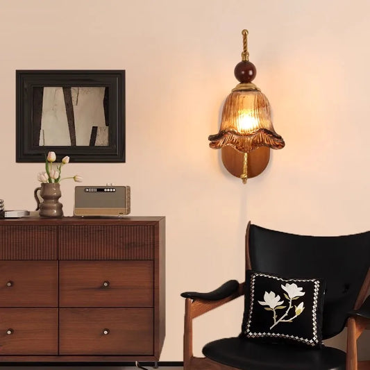 A vintage-style wall sconce with a floral-shaped amber glass shade and a gold arm mounted on a wooden oval backplate, shown in a bedroom setting.