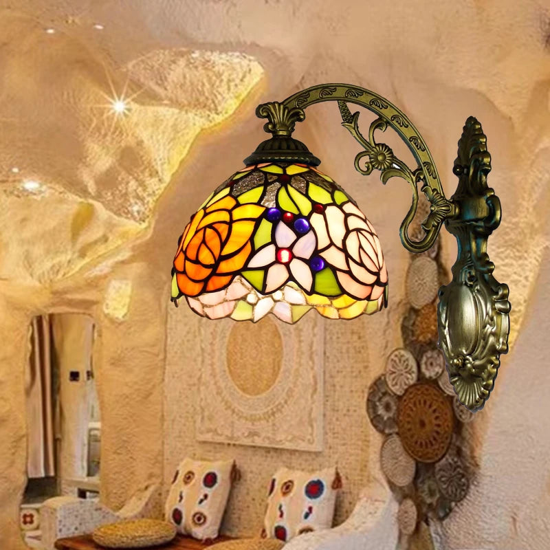 Tiffany style wall sconce with a stained glass shade featuring a rose and floral design, mounted on an ornate bronze-finished arm in a room setting.