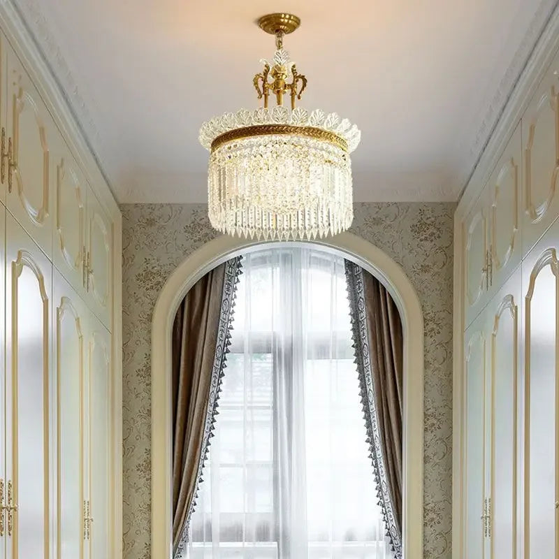 A gold and crystal tiered chandelier with cascading teardrop crystals, shown hanging in a hallway with arched doorways and built-in cabinets.