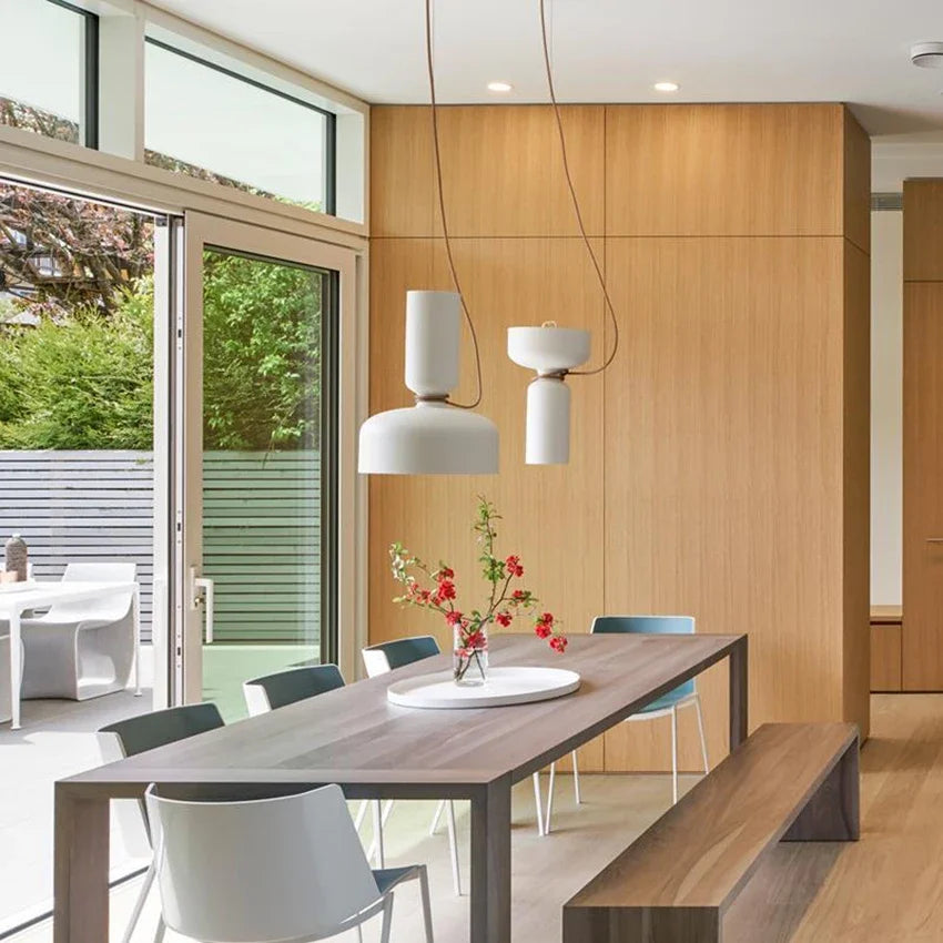 A dining room setting with two white pendant lights hanging above a wooden table, with natural light streaming in.
