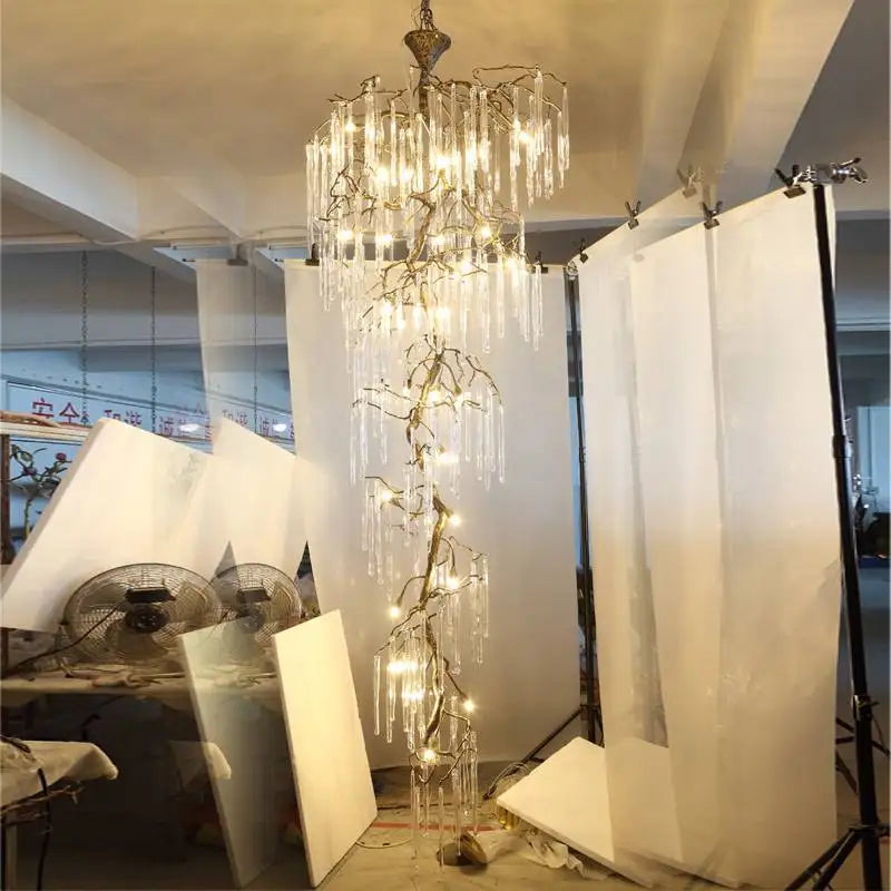 A tall, cascading chandelier with a gold branch-like frame and clear crystal droplets, shown in a studio setting with professional lighting equipment and backdrops.